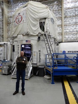 Photograph of Will Daniels standing in larger aircraft hanger with shiny, puffy insulated walls, in front of white, balloon-like space-capsule looking object that will be his home for 45 days.